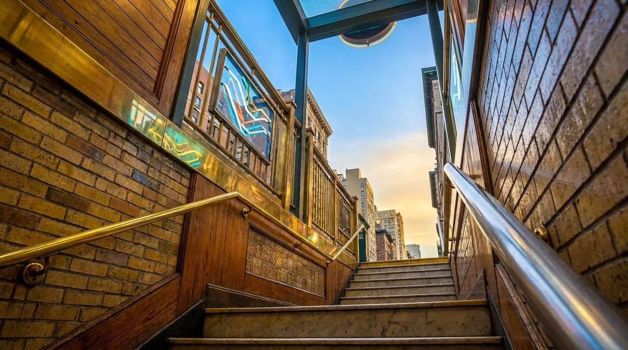 Bright dusk low-angle NYC subway stairwell on WC 2026 match night, brick wall, brass railings, polished steel handrail, neon-lit entrance opening to sky above, no people