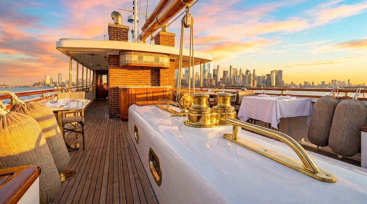 Bright sunny golden-hour low-angle view from NYC ferry deck on WC 2026 match day, polished white hull, brass deck fittings, NJ skyline ahead, vivid sky, no people