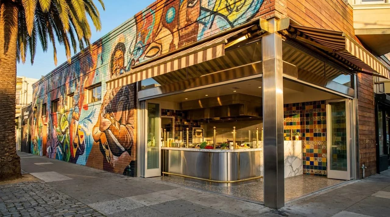 Bright sunny golden-hour low-angle SF Mission burrito shop corner on WC 2026 match day, colorful mural facade, polished steel counter visible through window, palm tree shadow, vivid sky, no people