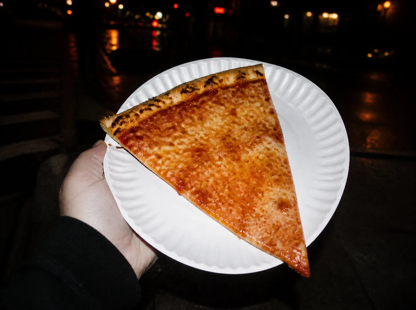 A single large plain cheese pizza slice on a thin paper plate held over a dark pavement at night, lit by a harsh direct camera flash with blurred streetlights behind