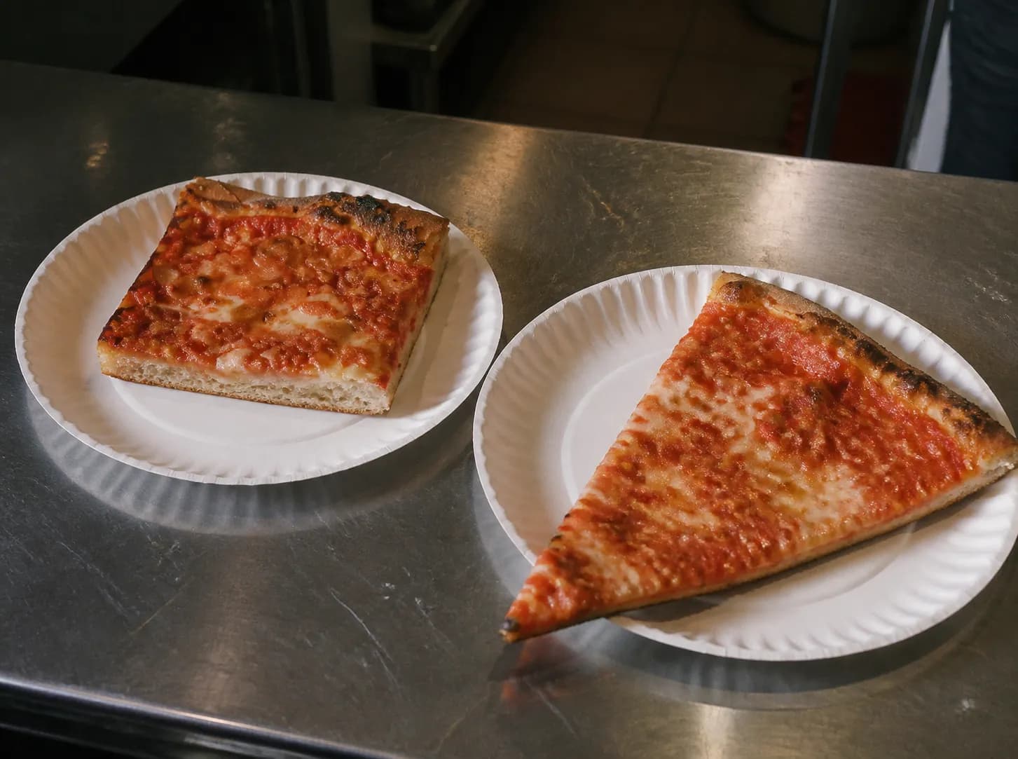 Two plain pizza slices on white paper plates on a scuffed steel counter in a small takeaway shop, one thick square focaccia-style slice and one thin wide triangular slice