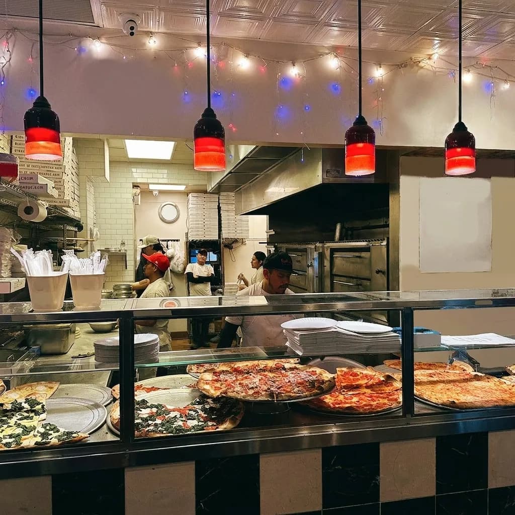 Late-night New York pizza counter with whole pies under warm red pendant lights