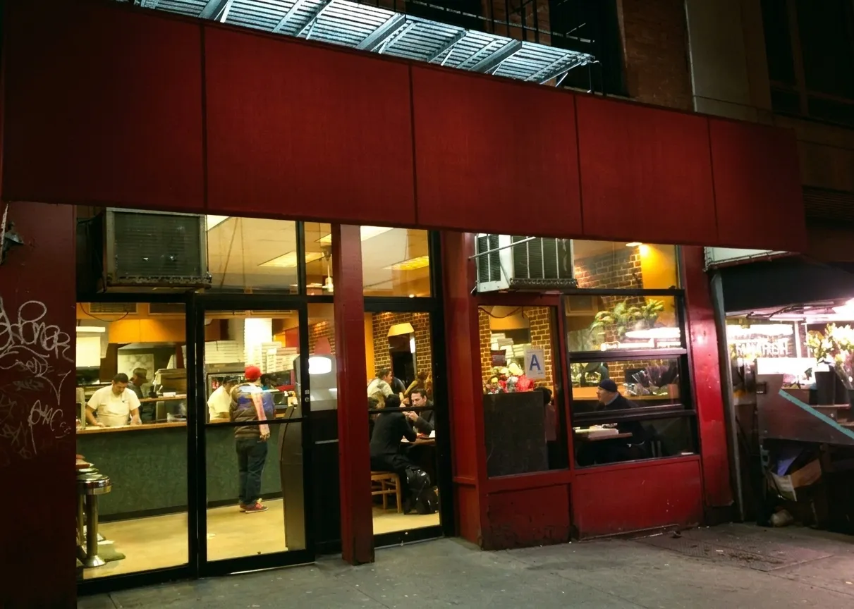 Corner pizza shop with a red awning glowing at night on upper Broadway in Manhattan