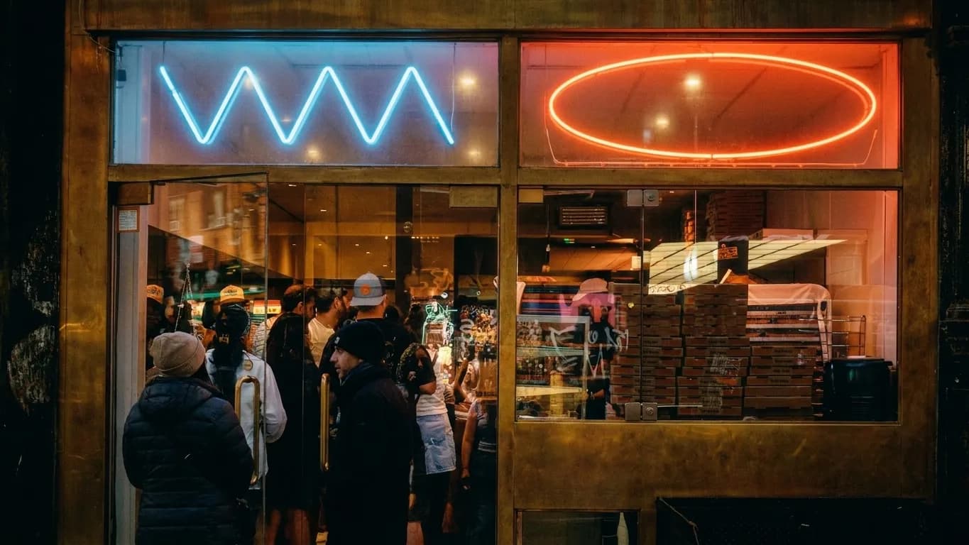 Neon-lit pizzeria storefront glowing on a dark Lower East Side street at night with a crowd inside