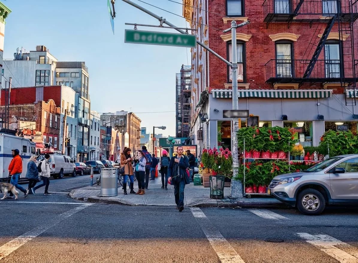 Wide view of a busy Williamsburg street corner with brick tenements, a flower stand and pedestrians crossing