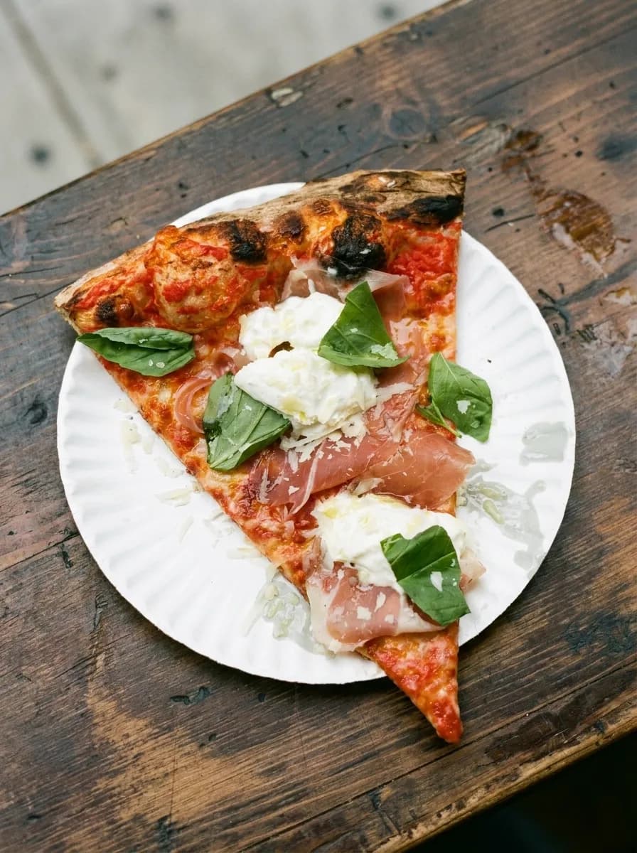 Overhead view of a burrata-topped New York pizza slice with basil on a white paper plate on a worn wooden counter in Williamsburg
