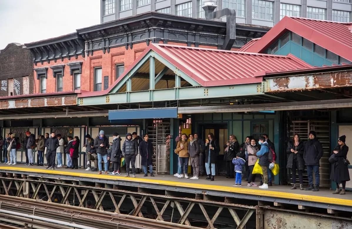 Commuters waiting on the elevated Marcy Avenue subway platform in Williamsburg with brick facades and red station roofs behind