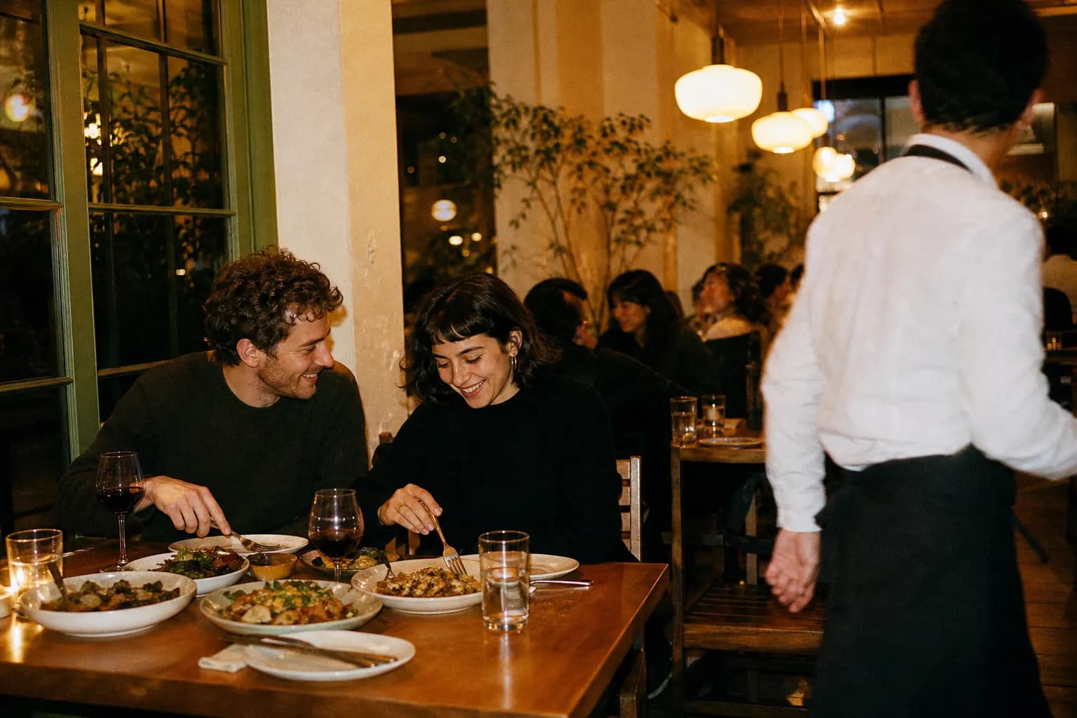 A cozy neighborhood restaurant interior in the early evening with warm pendant lights and diners sharing plates