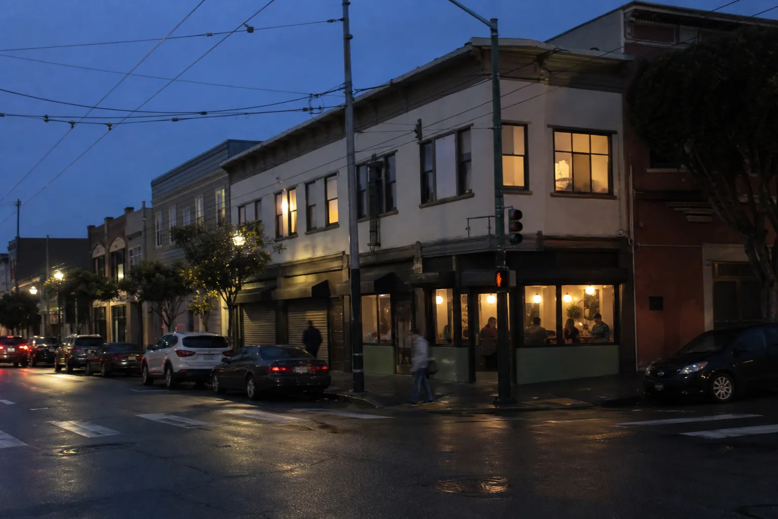 Dusk on a low-rise Mission District commercial block in San Francisco, wet asphalt and overhead Muni wires, warm light spilling from a corner restaurant window