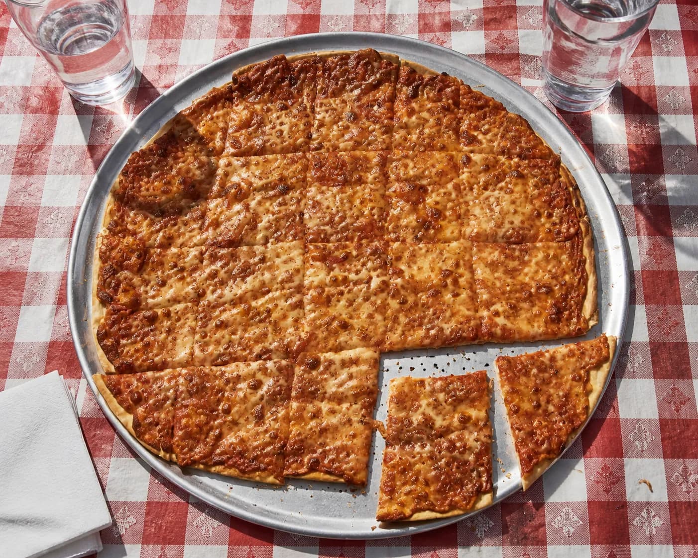 Overhead flat lay of a cracker-thin round pizza cut into small tavern squares on a perforated aluminium pan set on a red checkered tablecloth in hard midday light