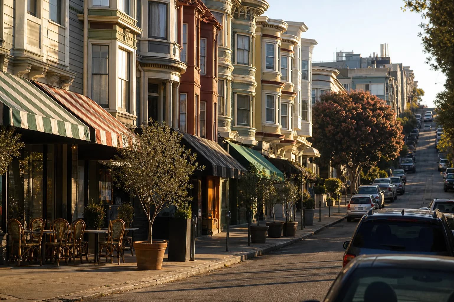 Late afternoon golden light on a narrow North Beach street in San Francisco, bay-windowed buildings with striped awnings, sidewalk cafe tables and potted olive trees