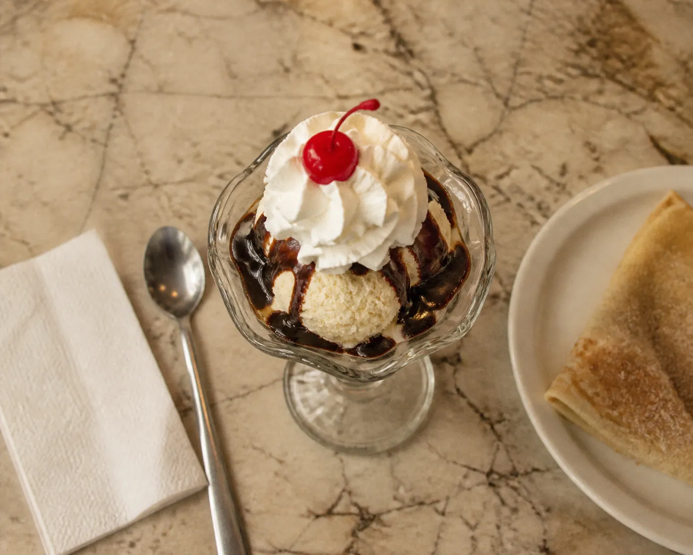 Overhead view of an old-fashioned ice cream sundae in a footed glass dish on a scuffed marble counter, with a long spoon, a paper napkin and a folded sugar crepe on a white plate at the edge of frame