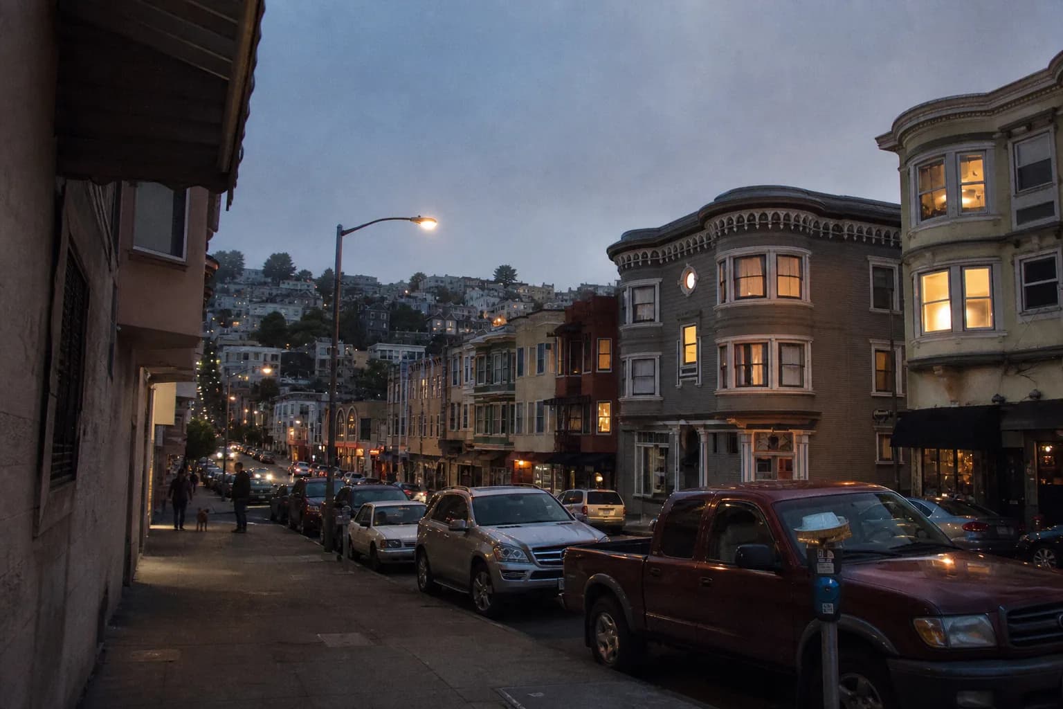 A narrow North Beach street in San Francisco at dusk with bay-windowed buildings, parked cars and hills glowing in the background
