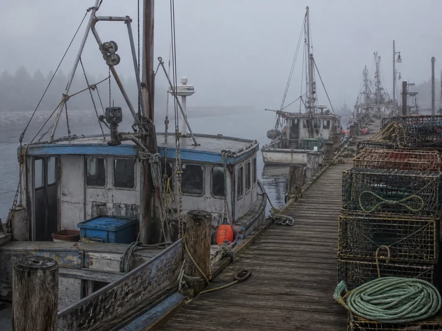 Wooden fishing boats moored along a foggy San Francisco pier with coiled rope and stacked crab pots on the dock