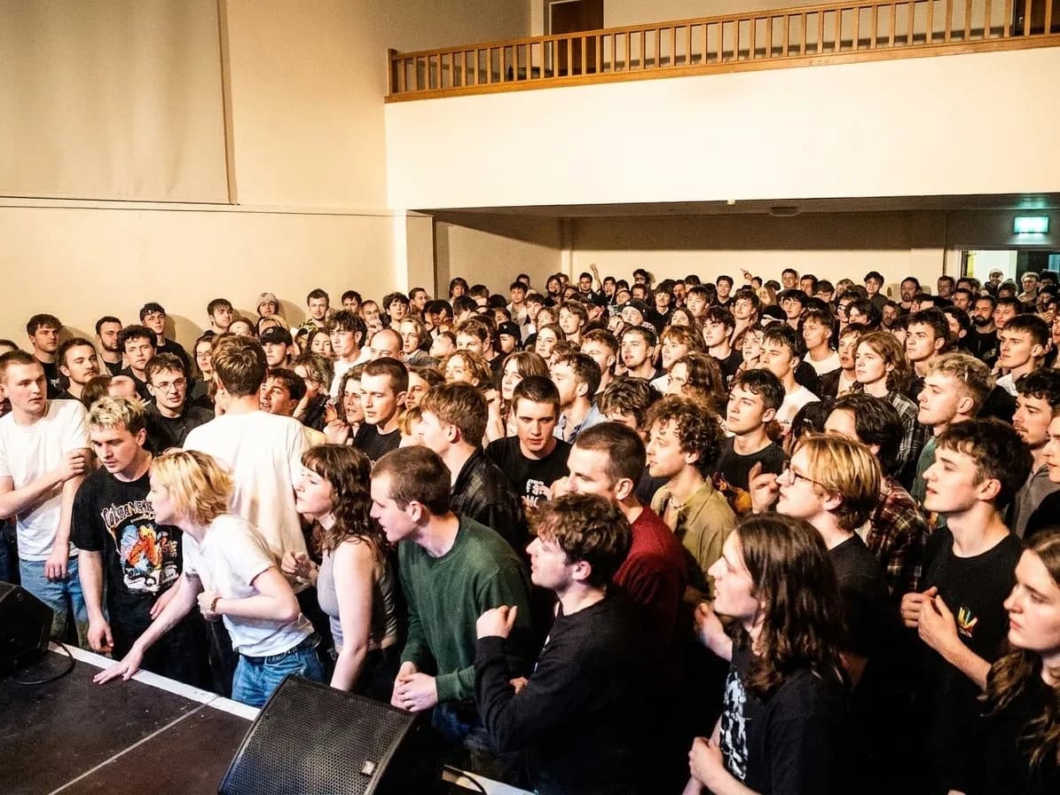 An all-ages gig in a small hall with a young crowd standing shoulder to shoulder facing a low stage under bright wash lighting