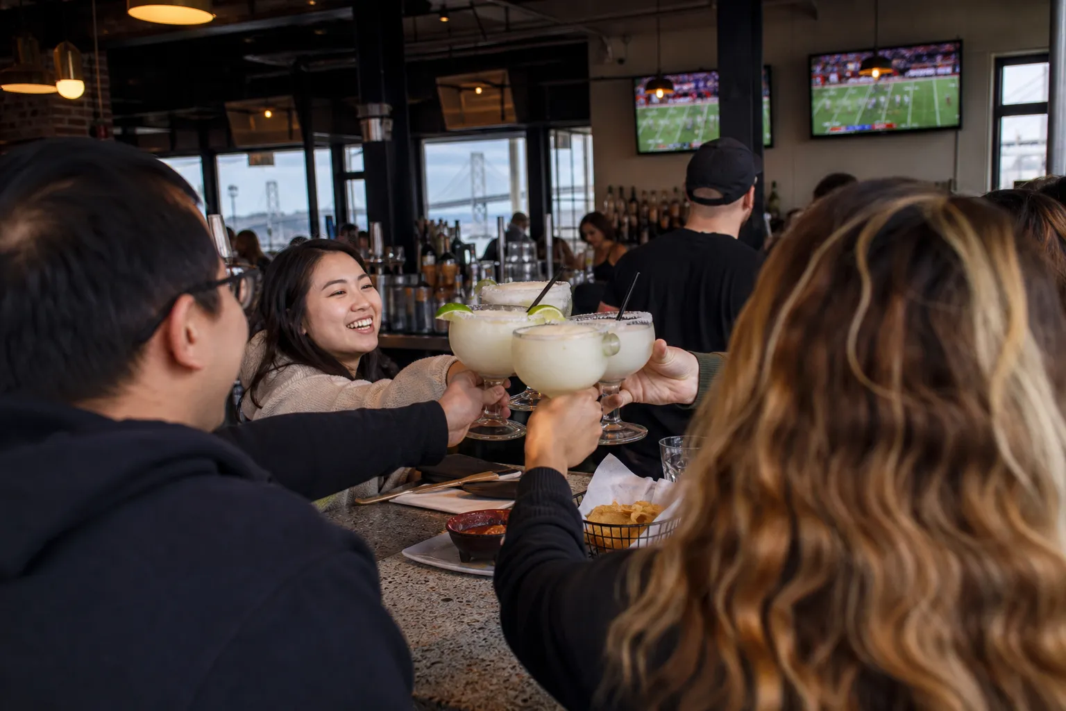 Friends clinking frozen margarita glasses over a wooden bar counter.