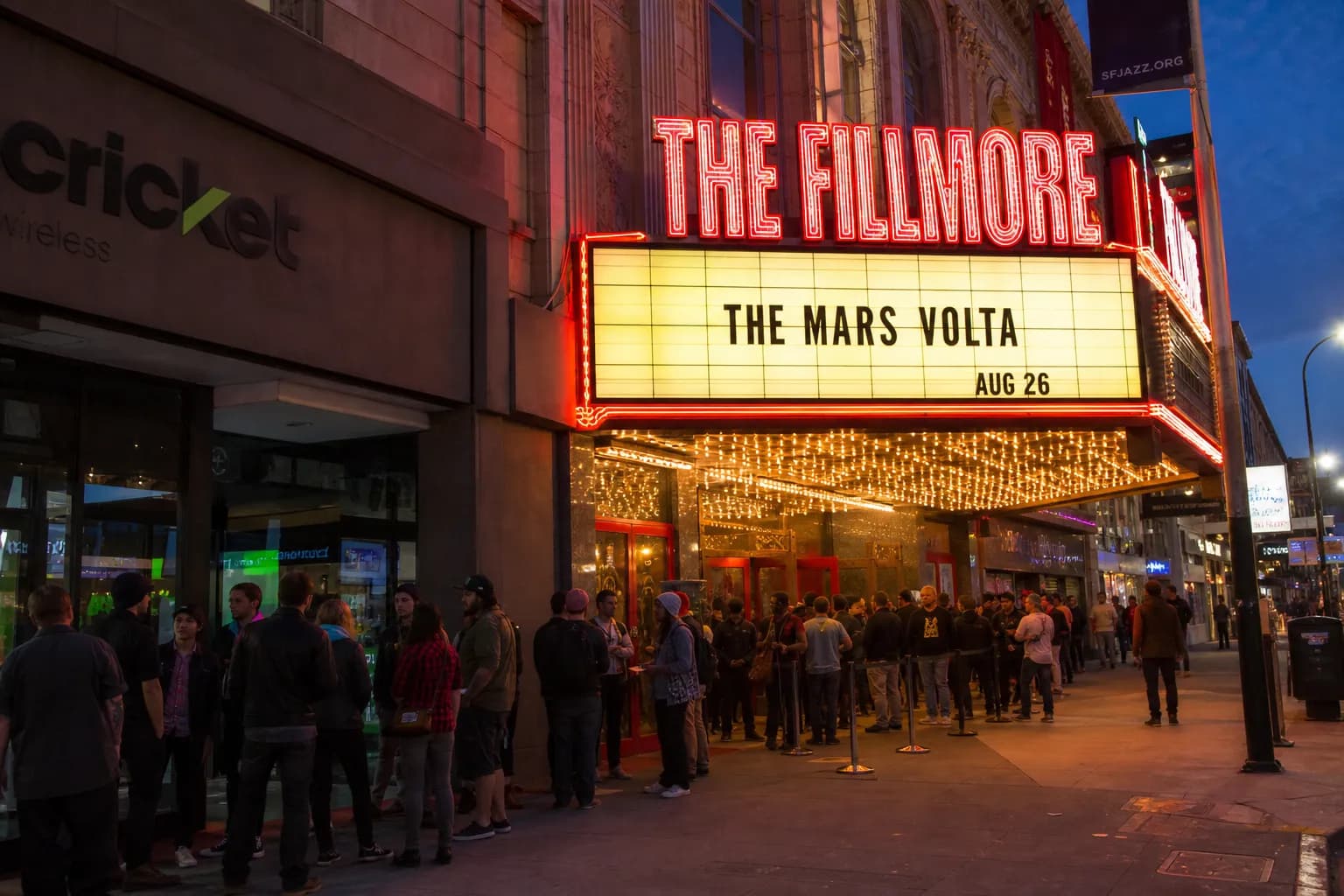 A wide shot of a historic concert venue facade in San Francisco at dusk with glowing neon signs and music fans gathering outside.