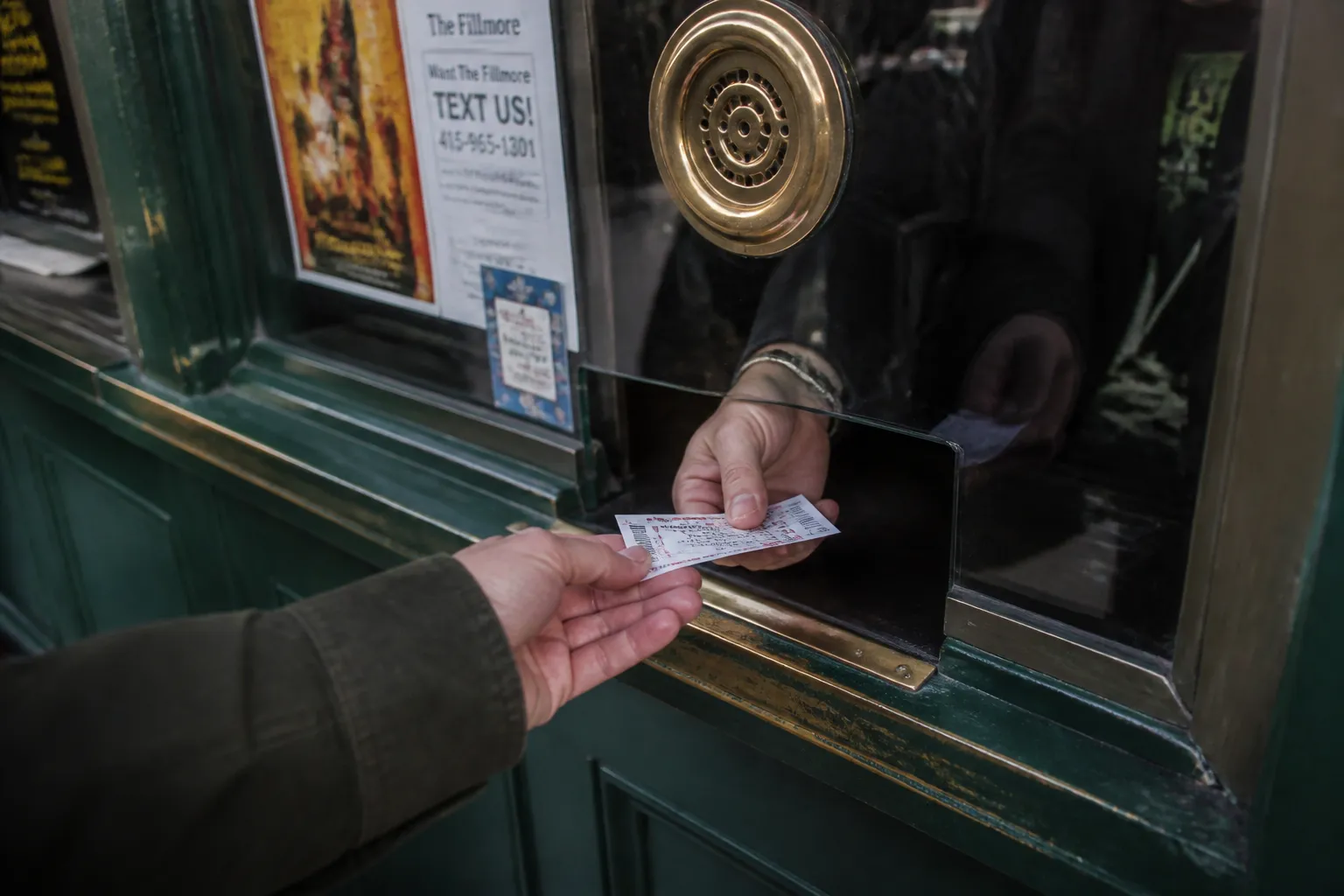 A close-up of a hand receiving a physical concert ticket from a vintage-style ticket booth window.