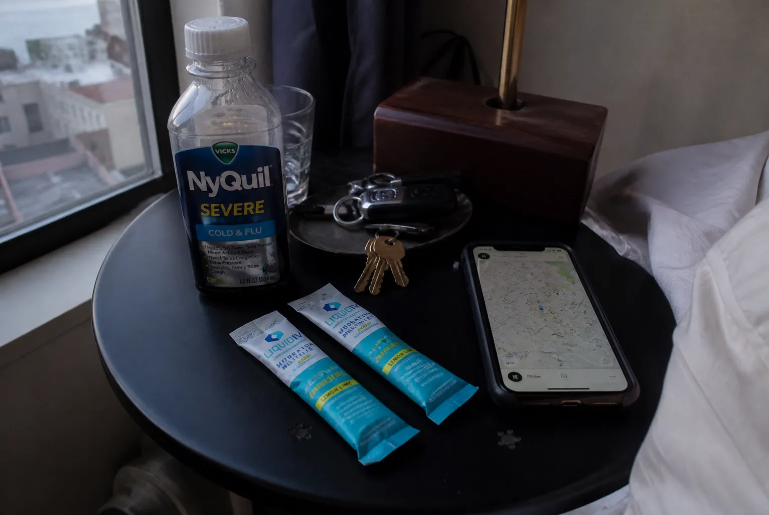 A minimalist tabletop setup of emergency pharmacy essentials like hydration packets, pain relievers, and a house key on a wooden bedside nightstand in a San Francisco apartment.