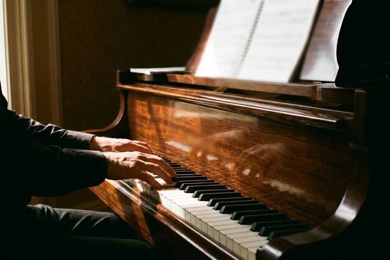 A pianist's hands on the keys of a grand piano seen from the side with blurred sheet music on the stand under warm directional light