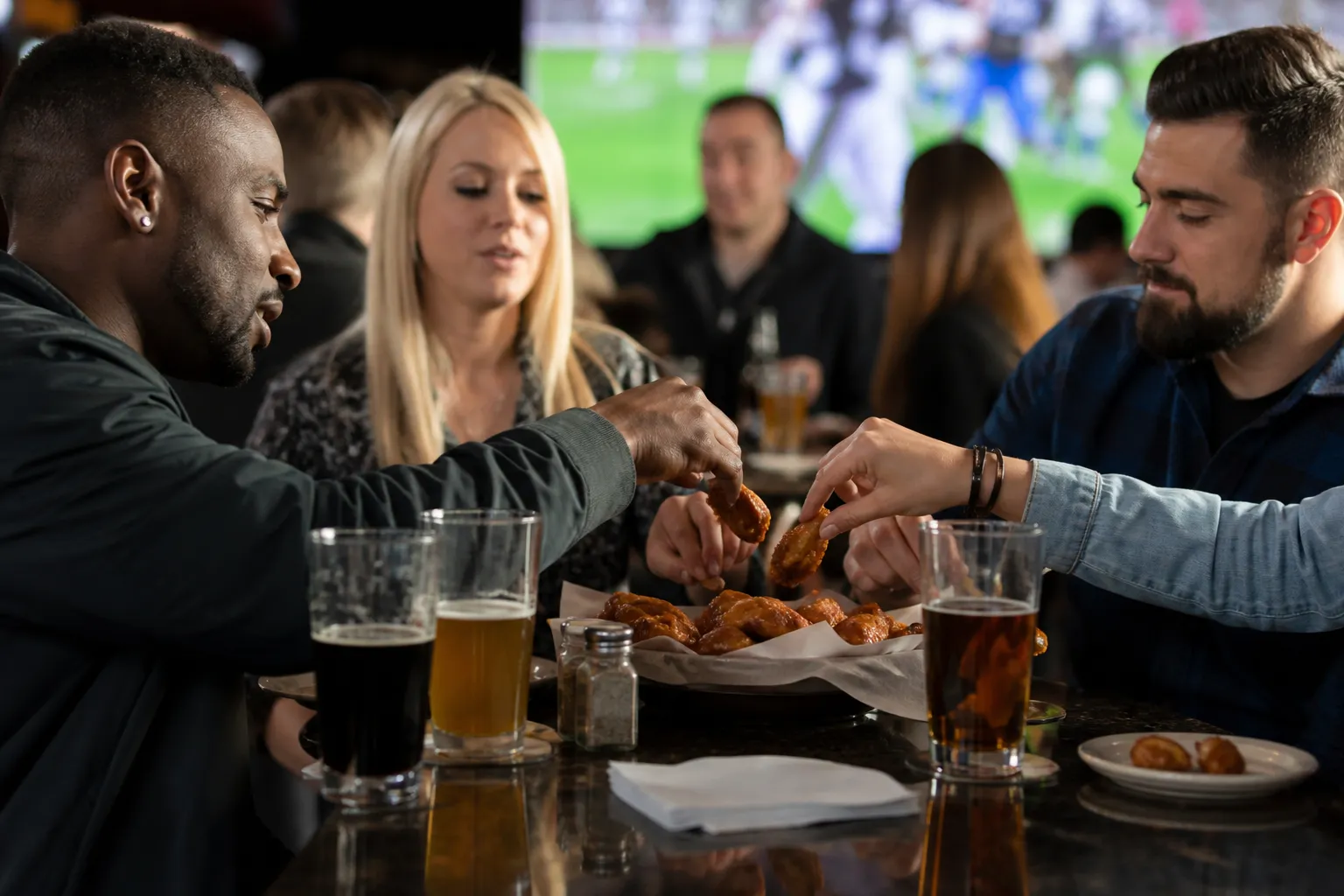 A group of friends sharing appetizers and drinks at a sports bar table with game screens visible in the background.