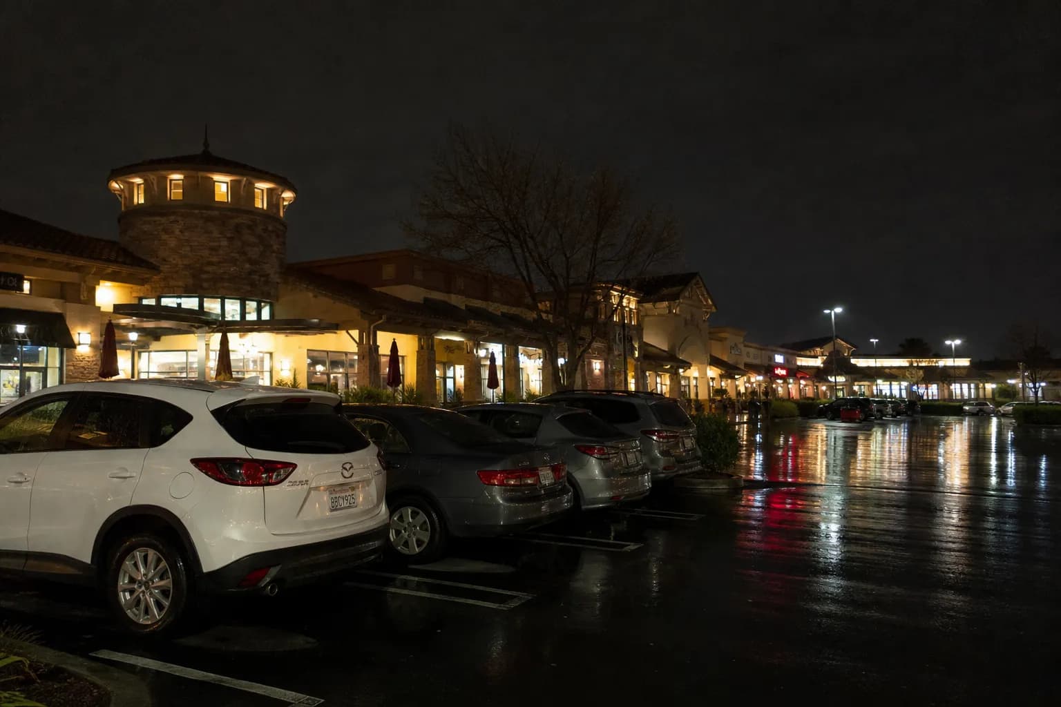 The warm glowing storefront lights of the Concord shopping plaza at dusk, reflecting off damp asphalt with cars parked outside.