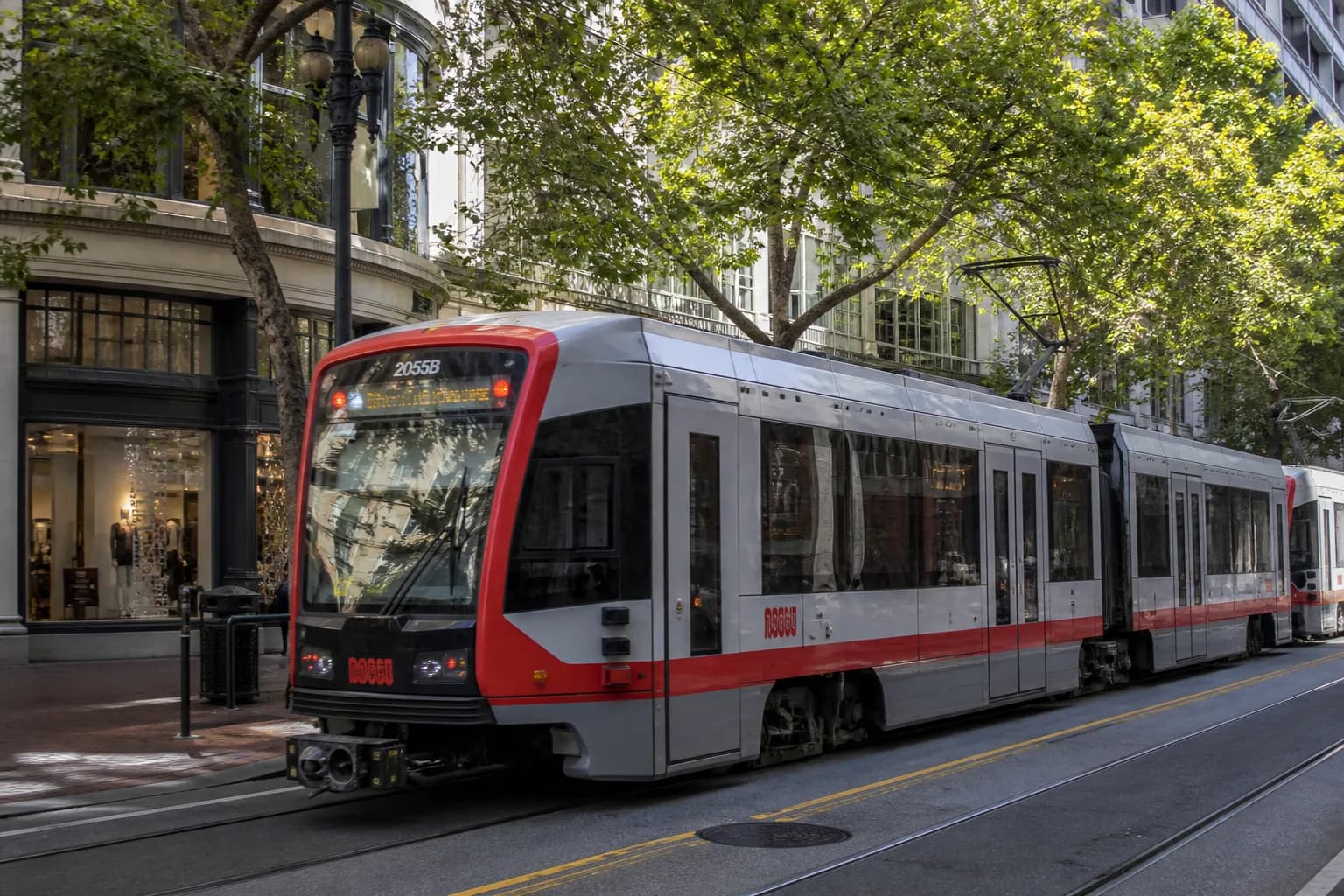 A modern San Francisco Muni light rail train passing through a busy shopping district with storefronts in the background under a bright California sky.