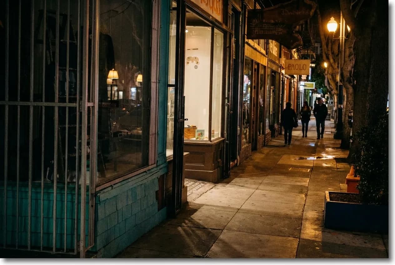 Narrow Hayes Valley side street at dusk, warm yellow light spilling from a small storefront window onto the sidewalk, a single person walking past darkened neighbouring shopfronts, streetlamp glowing overhead against a deep blue evening sky.