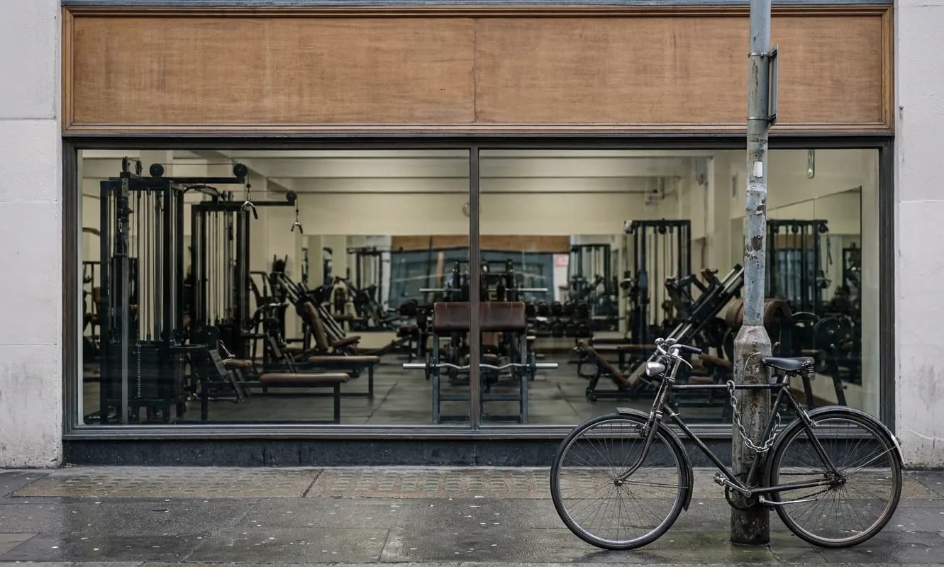 A ground-floor gym seen from the pavement through a wide window with rows of equipment inside and a blank unlettered fascia above