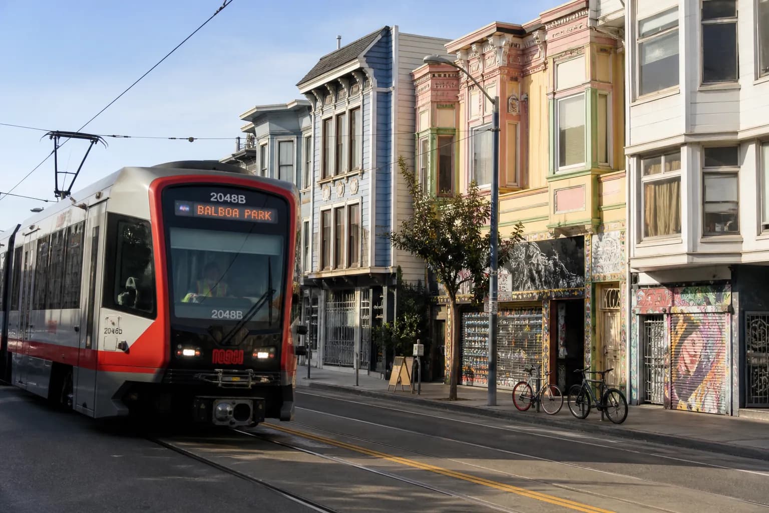 A classic San Francisco Muni train moving past vibrant storefronts on a sunny neighborhood street.