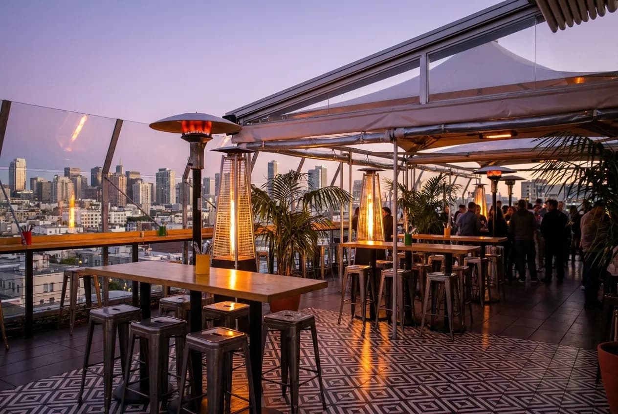 Covered rooftop bar terrace at dusk with glass walls, city skyline views, high-top tables, palm trees, and heat lamps glowing in San Francisco Mission District.