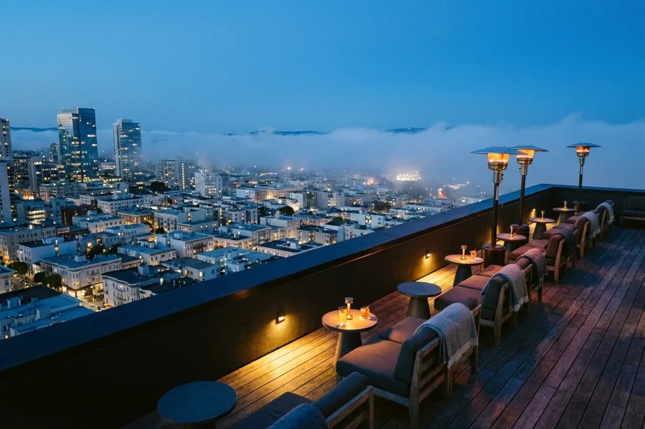 A San Francisco rooftop bar terrace at blue hour, low bench seating and small round tables along the parapet under glowing patio heaters, wool blankets on the seat backs, and a fog-hazed downtown skyline below