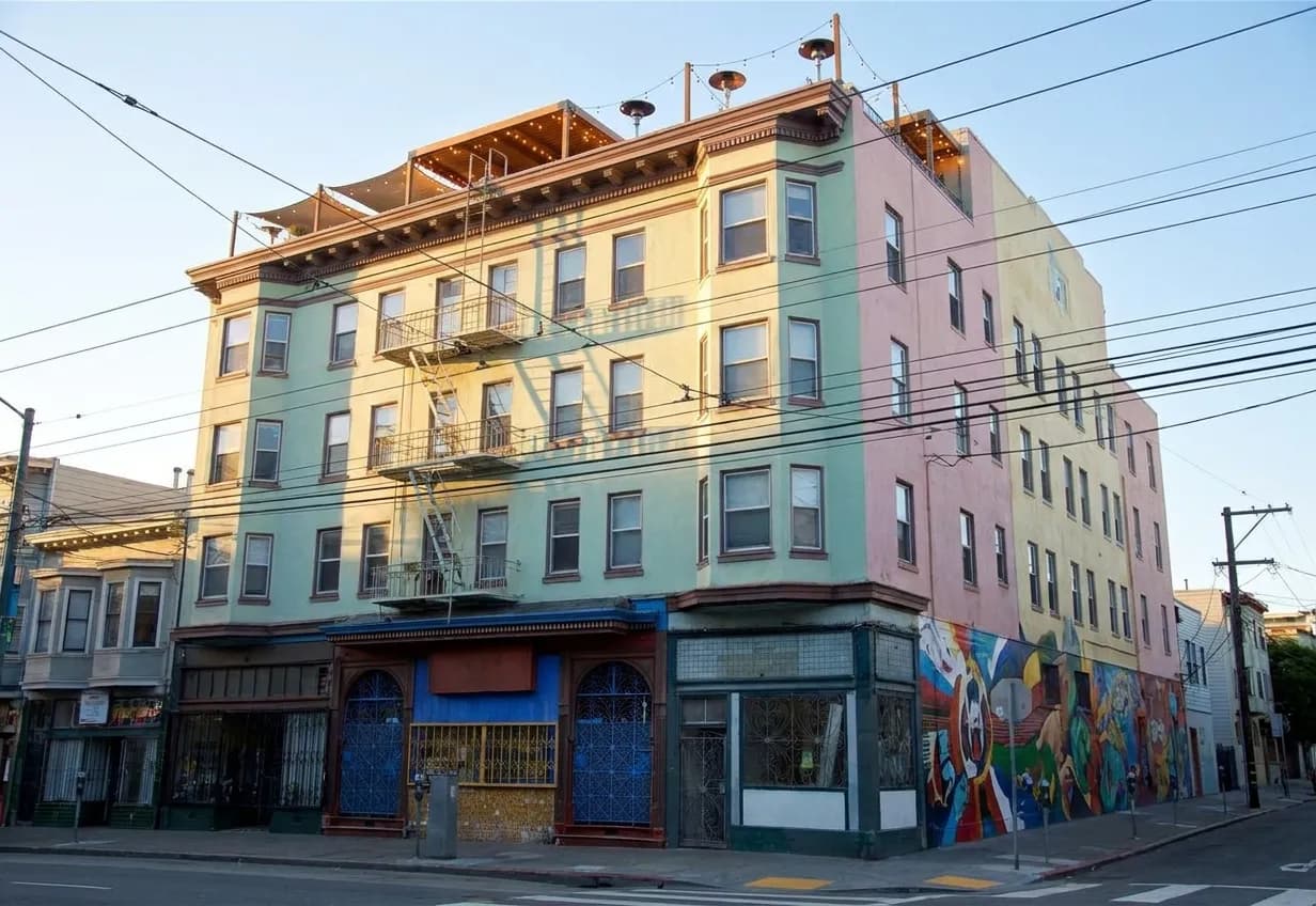 Late afternoon on Mission Street in San Francisco, looking up at a six-storey corner building with string lights and heat lamps visible over its rooftop parapet above a pastel stucco facade and a painted side-wall mural