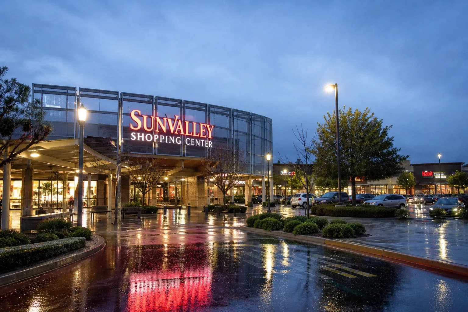 The modern glass and steel exterior entrance of the Sunvalley Shopping Center in Concord at dusk, illuminated with bright neon signage.