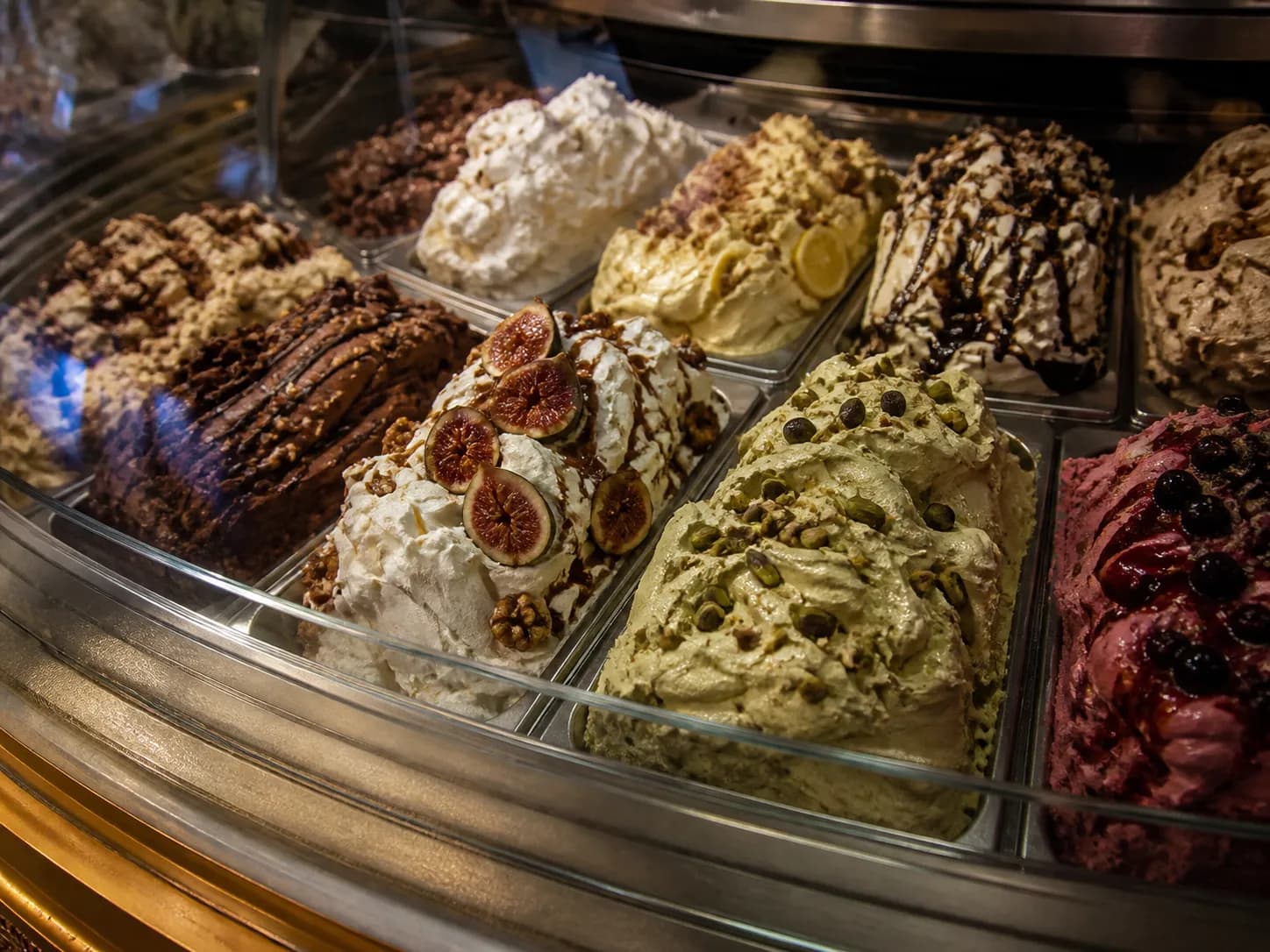 Artisan gelato mounded in stainless steel pans behind curved display glass, including pistachio, fig, chocolate and sour cherry