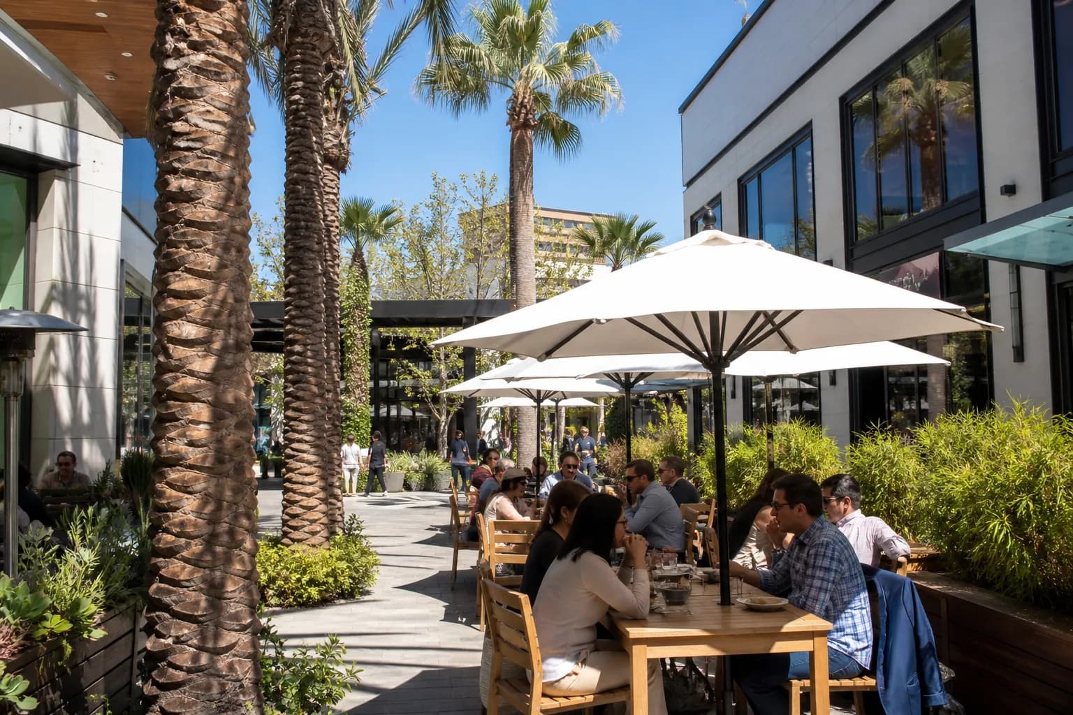 Sunny outdoor shopping plaza patio with dining tables under white umbrellas.
