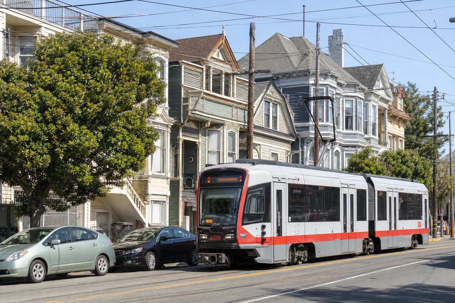 A classic red-and-white San Francisco Muni light rail vehicle traveling down a sunny city street with historic Victorian houses and green trees in the background.