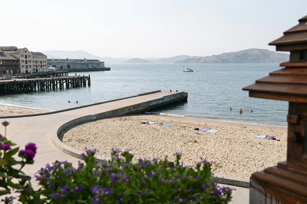 A small sheltered city beach on San Francisco Bay in early afternoon, coarse pale sand backed by a curved concrete promenade and timber piers, with flat water and hazy hills across the bay