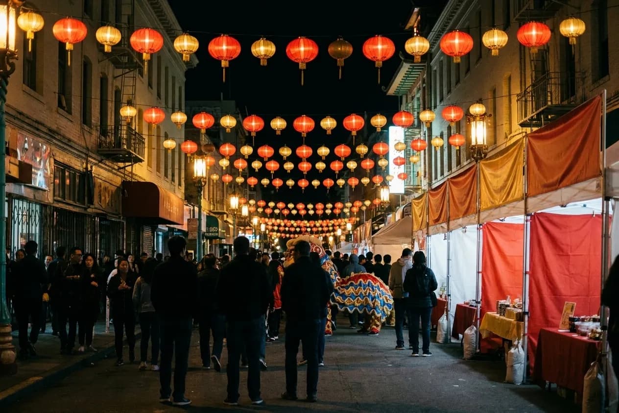 A Chinatown street in San Francisco closed for a festival evening, crowds beneath strings of red and gold lanterns with temporary stalls along one side and a lion-dance tail visible in the throng