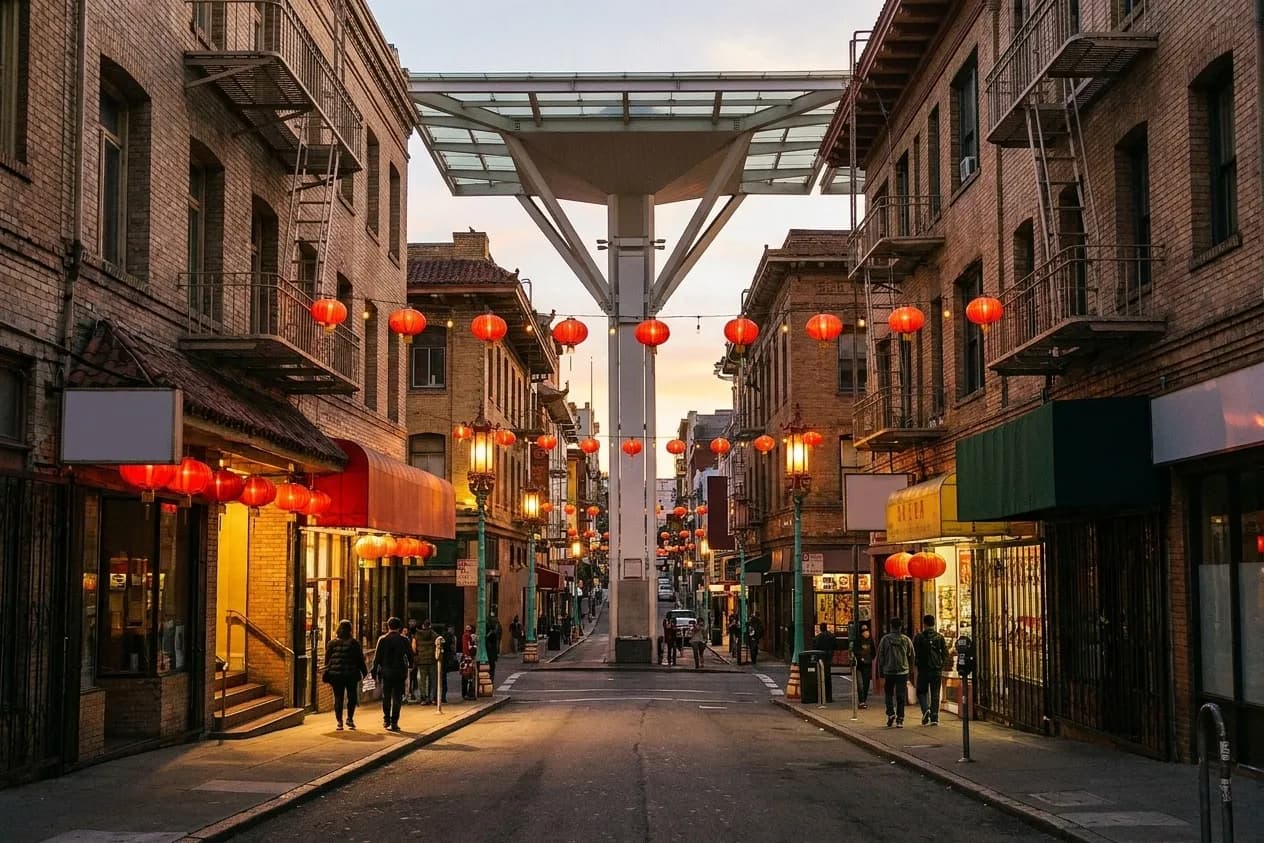 A narrow sloping Chinatown street in San Francisco at dusk with red lanterns strung overhead and fire escapes on the facades
