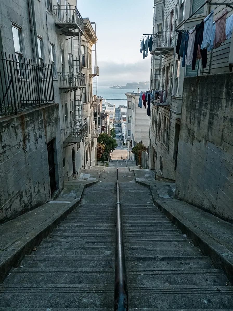 A flight of concrete public steps climbing between retaining walls and narrow buildings, with fire escapes above and a slot of bay view at the top