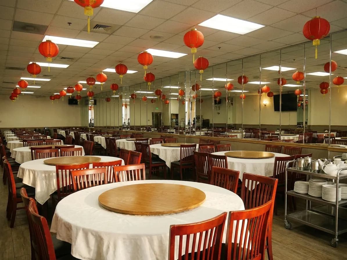 A large Chinese banquet dining room with round white-clothed tables and lazy susans, red-backed chairs and hanging lanterns overhead