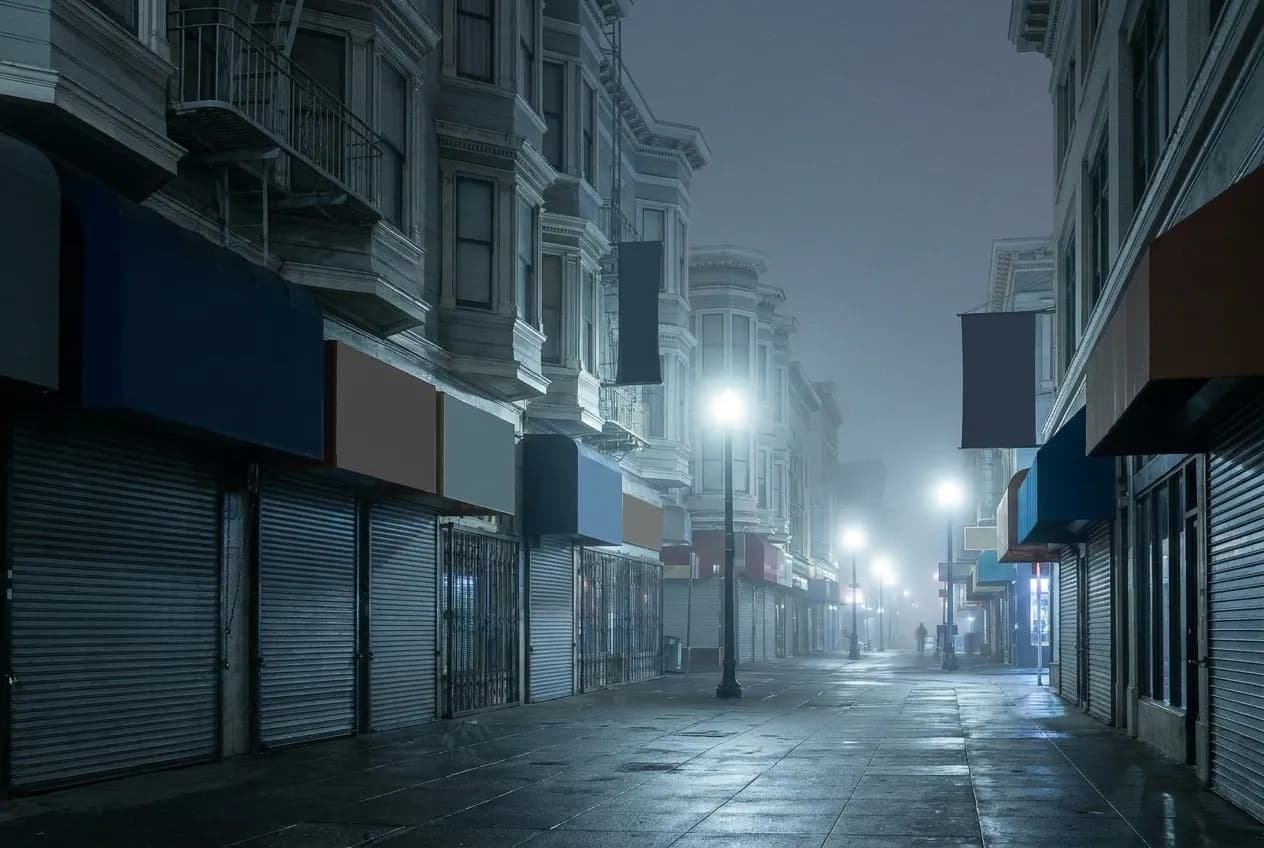 A commercial street after closing, shutters down and the pavement empty under streetlights