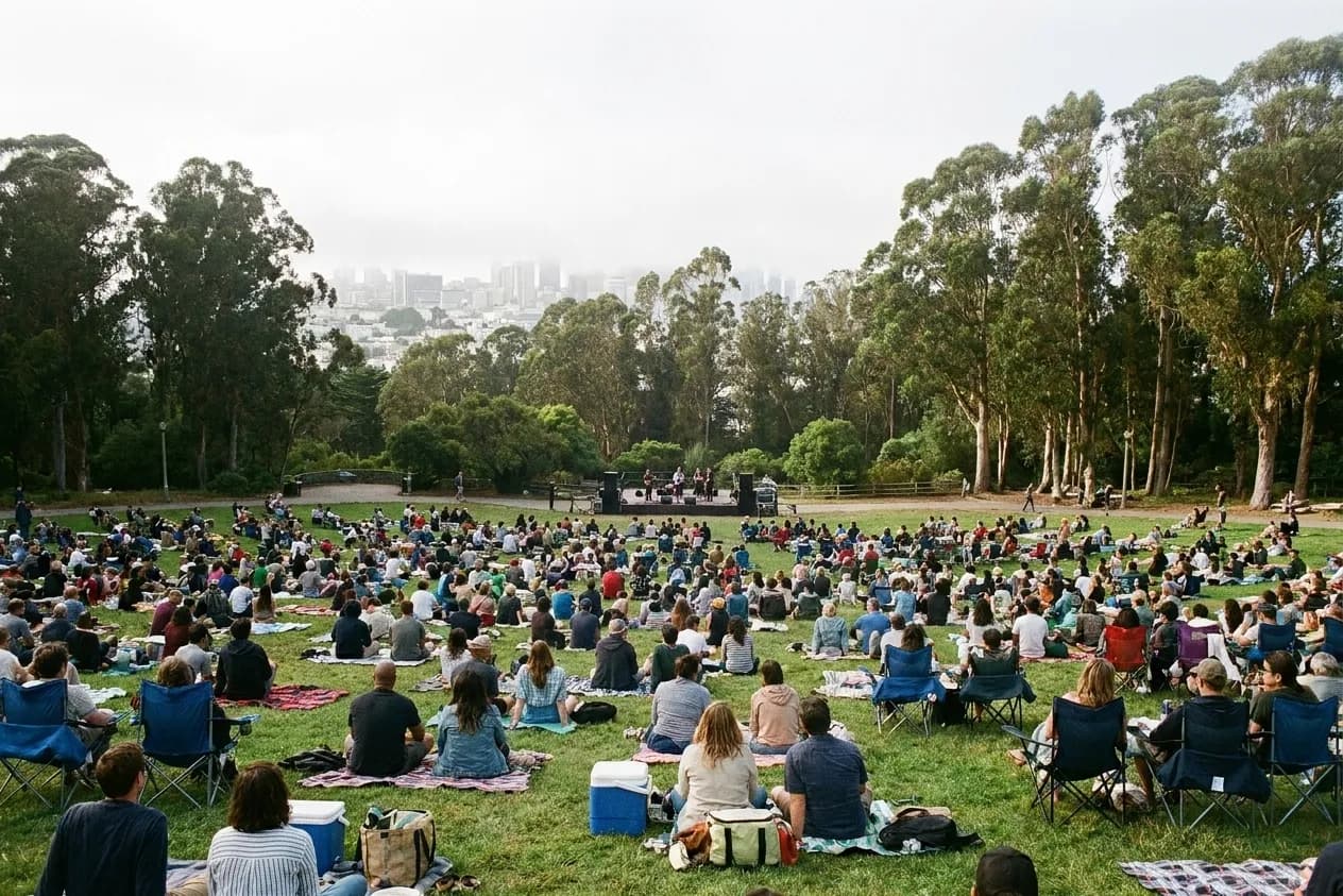 A free outdoor concert in a eucalyptus-ringed San Francisco meadow, hundreds of people seated on blankets down a grassy slope toward a small stage under high coastal fog