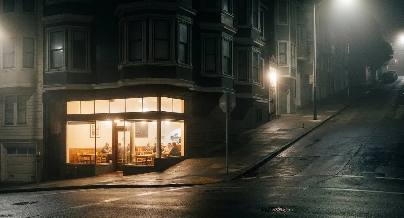 A lit San Francisco restaurant window at night on a sloping street with dark Victorian upper floors above and damp reflective pavement