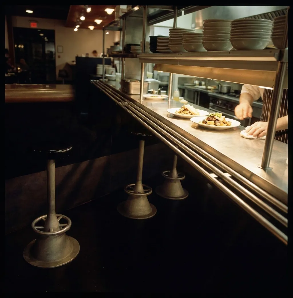 A stainless kitchen pass at the end of service in San Francisco with two plates under a warm lamp and a dimmed dining room behind