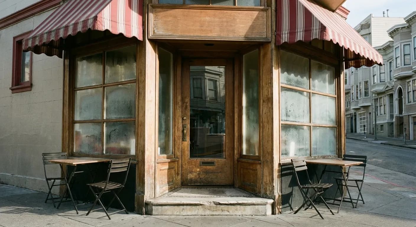 The chamfered corner entrance of a North Beach cafe on a bright cold afternoon, under a faded striped awning with a worn marble step and two empty pavement tables, a steep street rising behind