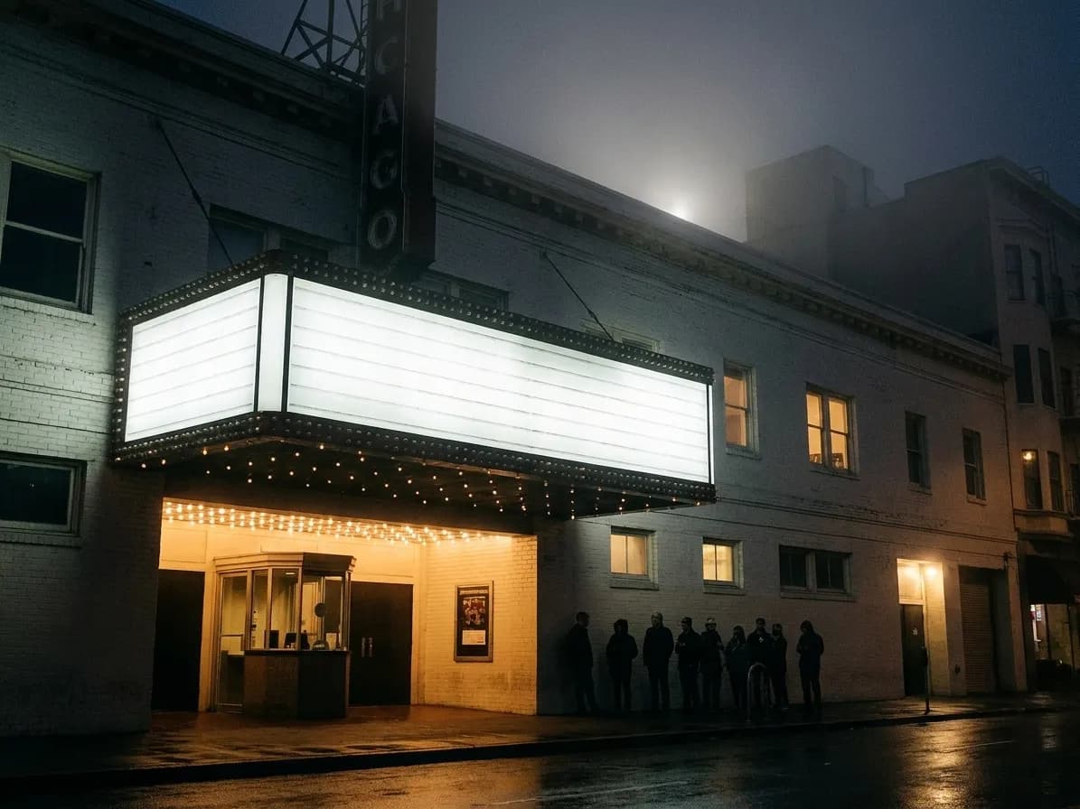 The entrance of an old San Francisco music venue at night, a projecting marquee of unlit bulbs and blank panels above a glass ticket booth, with a silhouetted queue along the painted brick wall