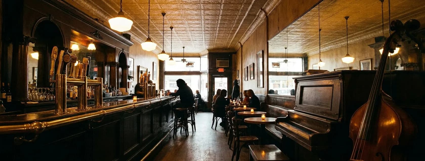 The interior of a small old San Francisco bar with an upright piano against a mirrored back wall and a double bass leaning beside it, warm amber light over candlelit tables and a brass-fitted bar
