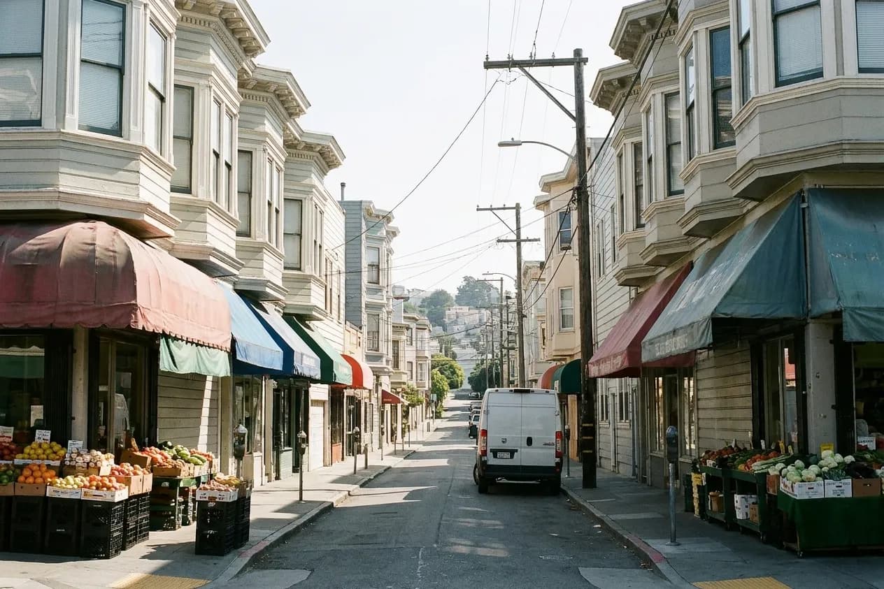A narrow San Francisco street of two-storey bay-windowed buildings with small shopfronts, faded awnings and produce crates on the sidewalk
